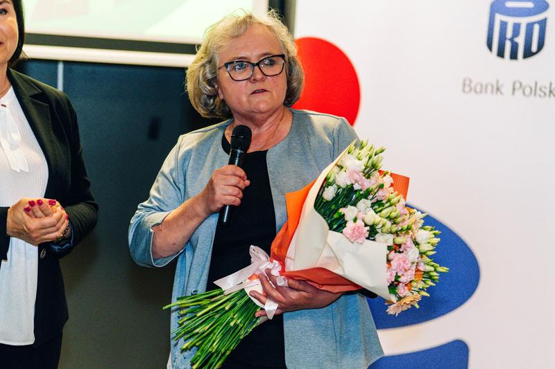 Uczestniczka rozdania wyróżnień PKO z bukietem kwiatów, mówiąca do mikrofonu w piwnicy hotelu.