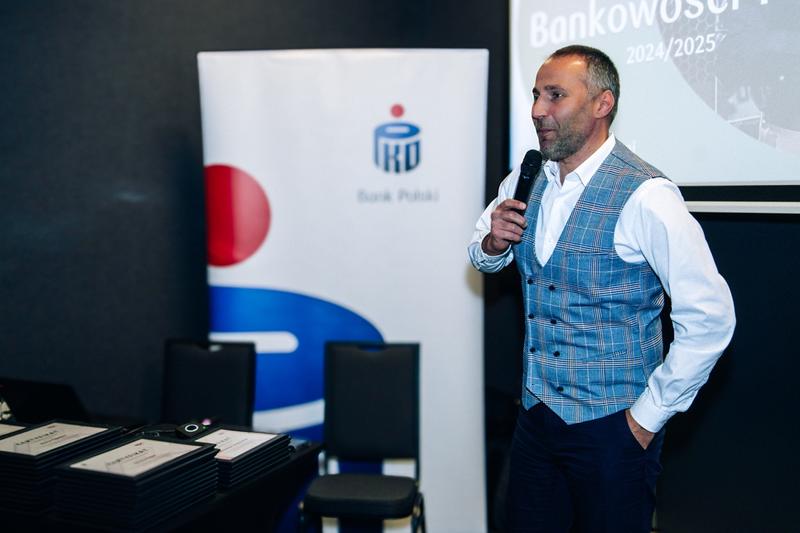 Mężczyzna przemawia podczas rozdania wyróżnień w hotelowej piwnicy, w tle certyfikaty PKO.