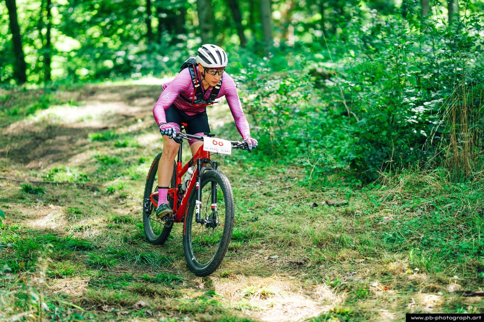 Zawodniczka MTB w różowym stroju jedzie przez zielony las podczas wyścigu.