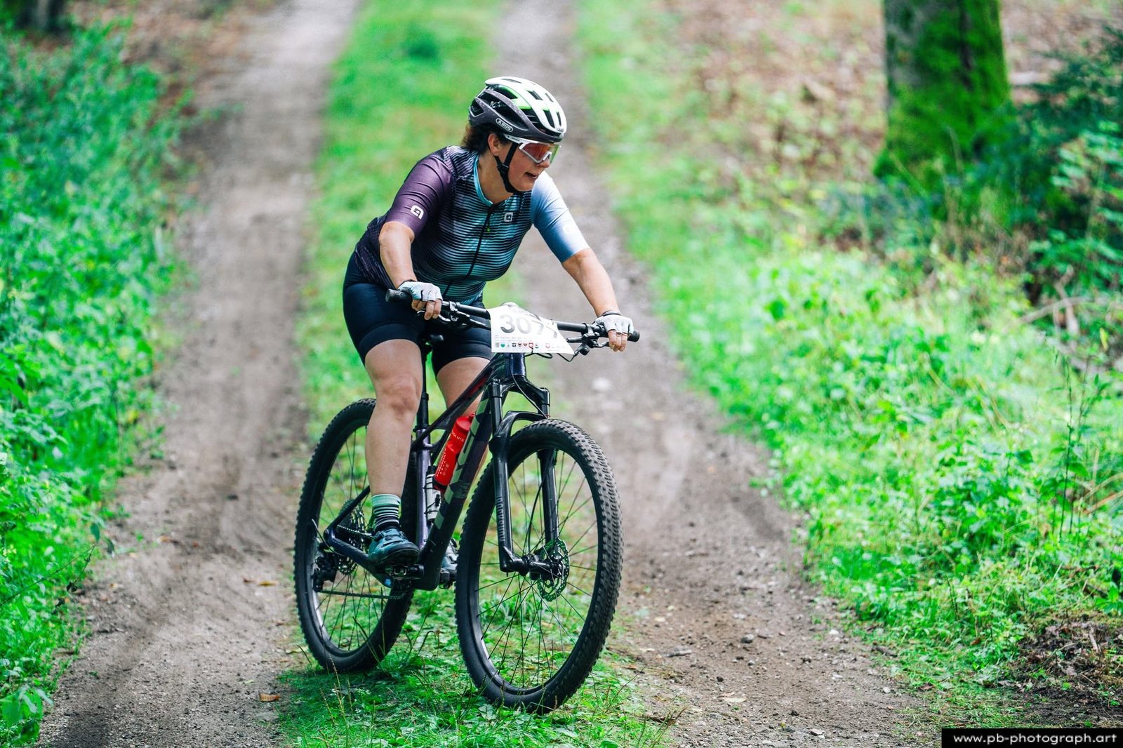 Zawodniczka MTB w ciemnym stroju jedzie szeroką leśną drogą podczas zawodów.