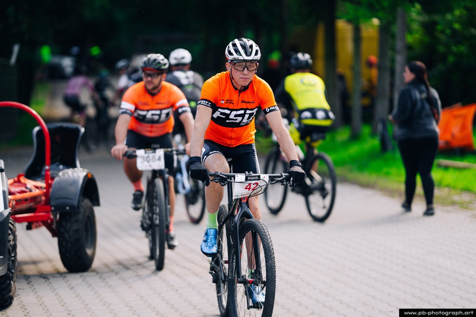 Zawodnik MTB w pomarańczowym stroju na rowerze, w tle inni uczestnicy wyścigu.